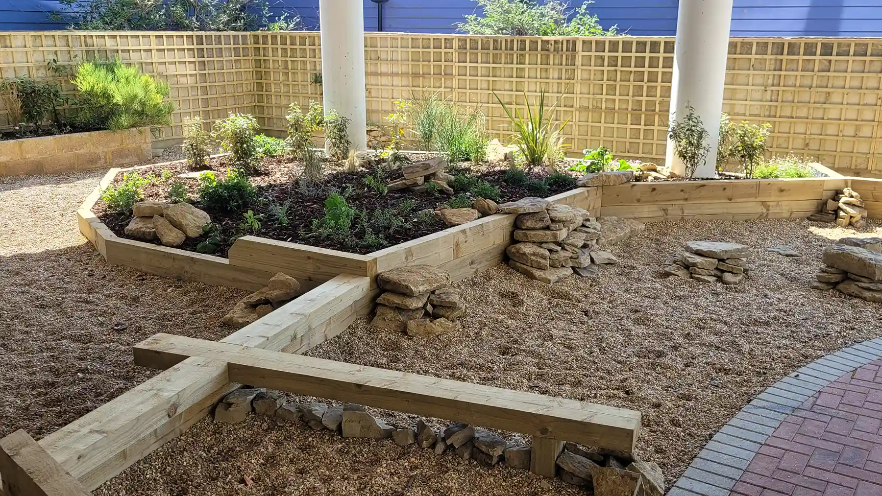 Shaded seated and raised planted bed made from softwood sleepers, surrounded with clusters of rocks.