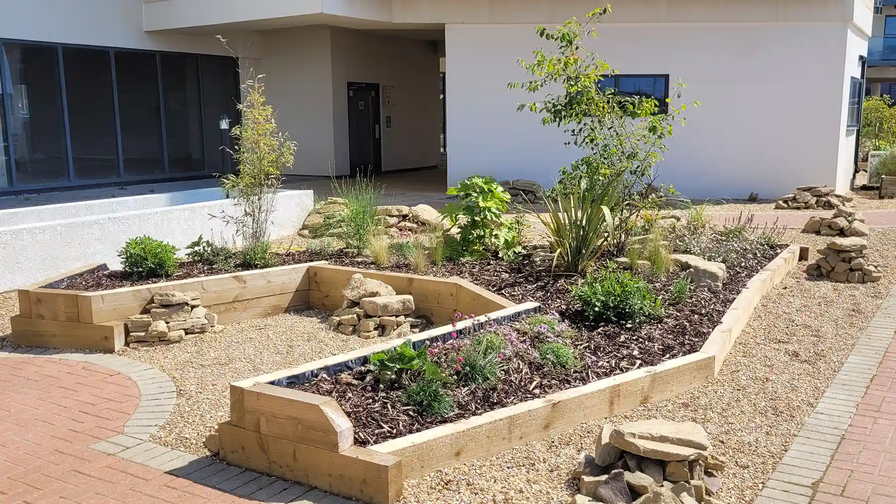 Angled raised sleeper flower bed surrounded with clusters of rocks and shingle.