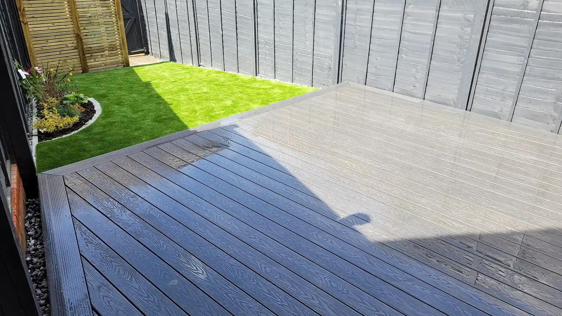 Grey composite decking with artificial grass and flower bed with screened of area for bins.