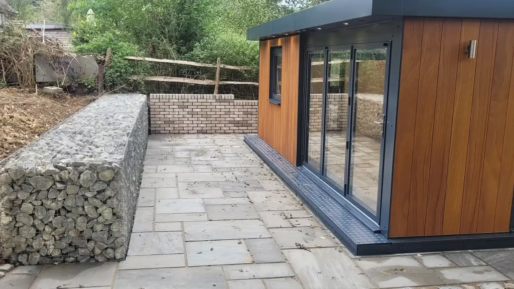 Gabion wall in front of a sandstone patio and garden building.