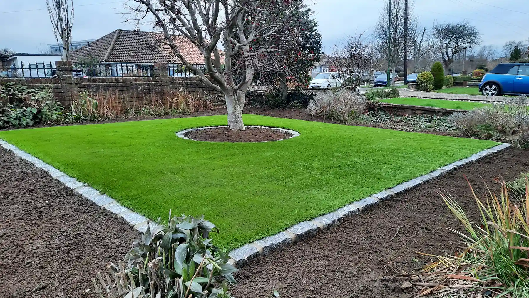 Front garden with artificial grass with acre tree in the centre with flower bed around the outside.