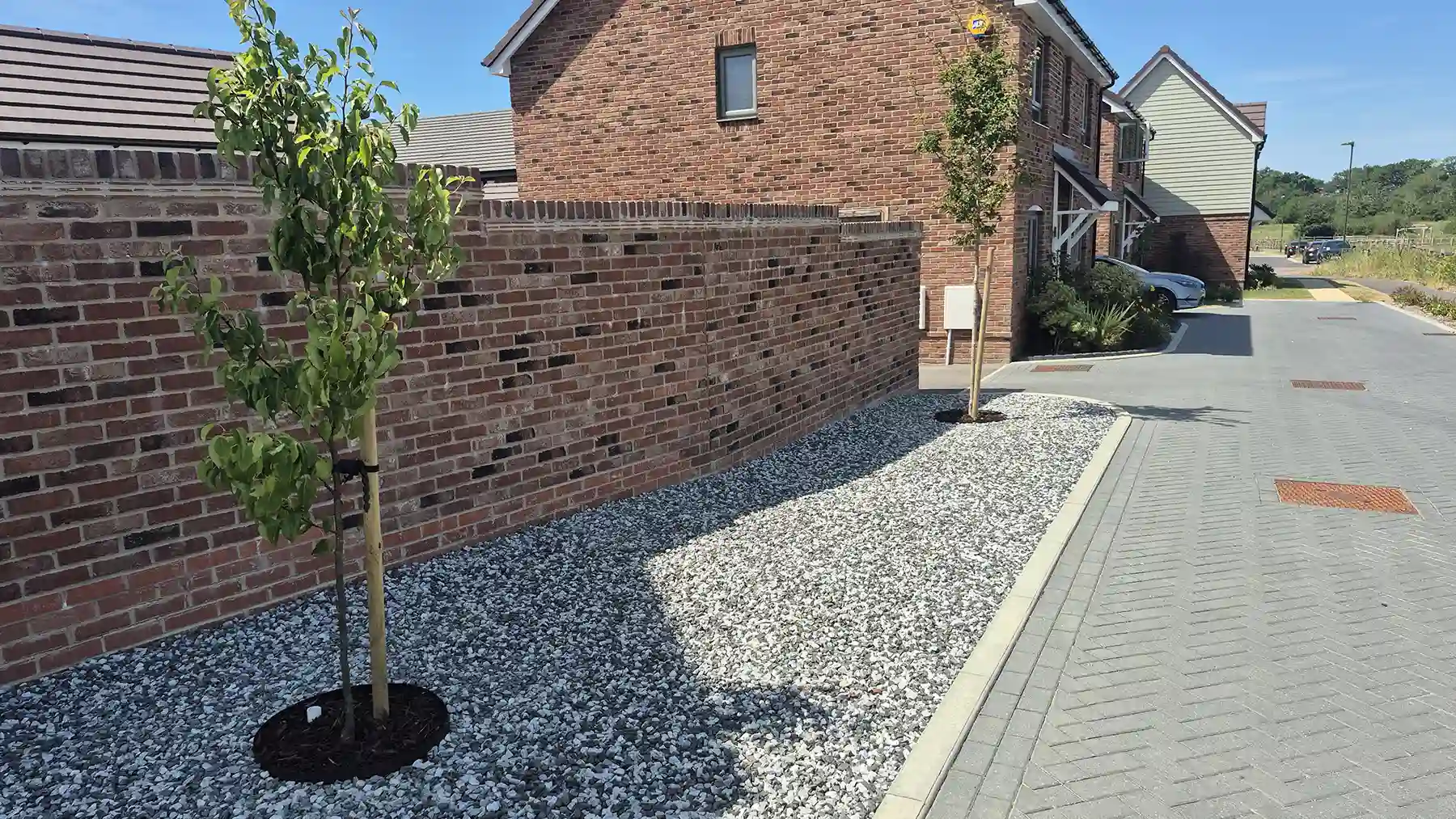 A landscaped front garden area filled with black ice aggregate stones, featuring a young staked tree growing alongside a high red brick boundary wall and paved driveway.