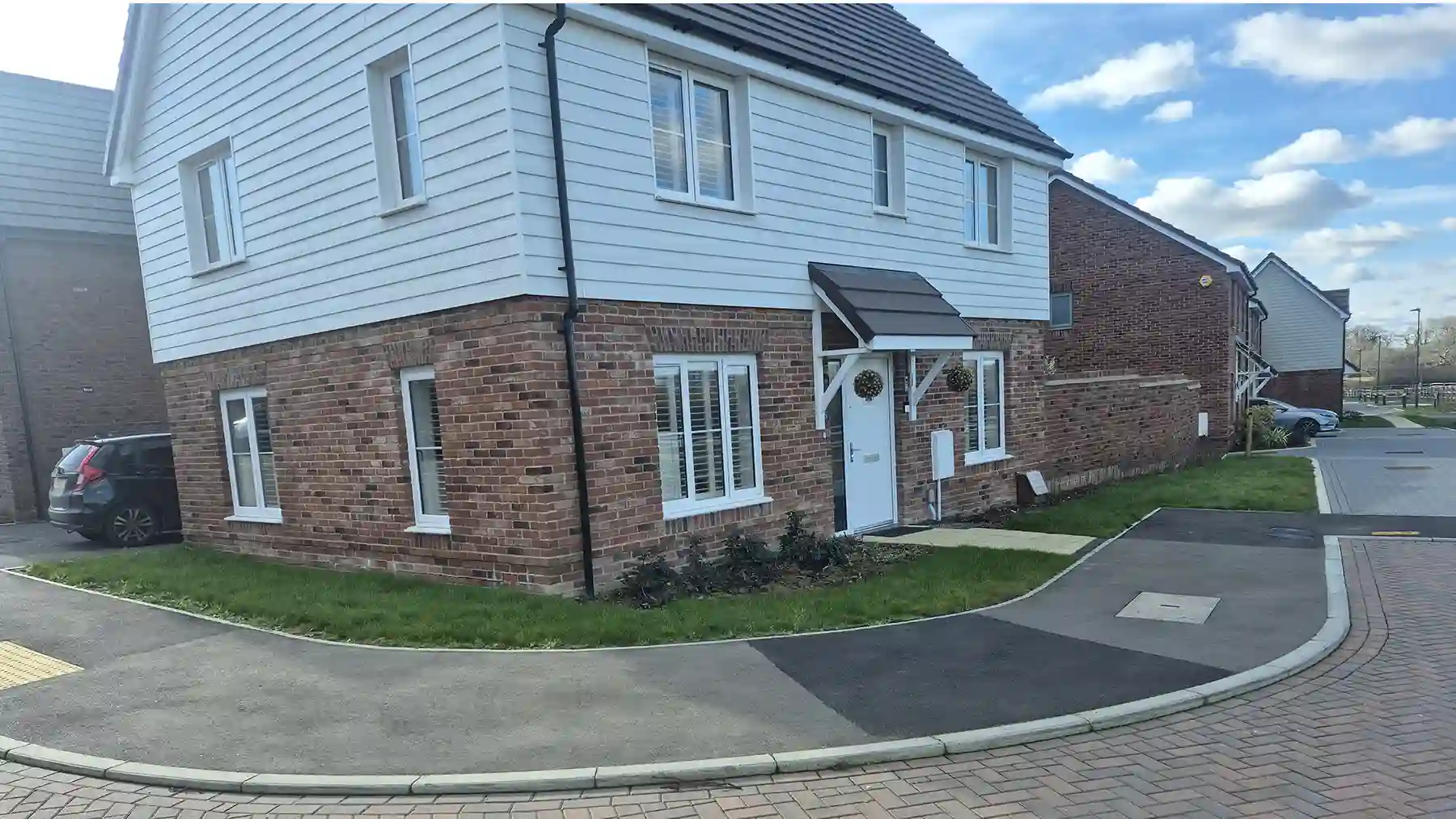 Wide view of a modern corner house with white cladding and red brick, surrounded by a green front lawn area bordering a paved pavement and street.