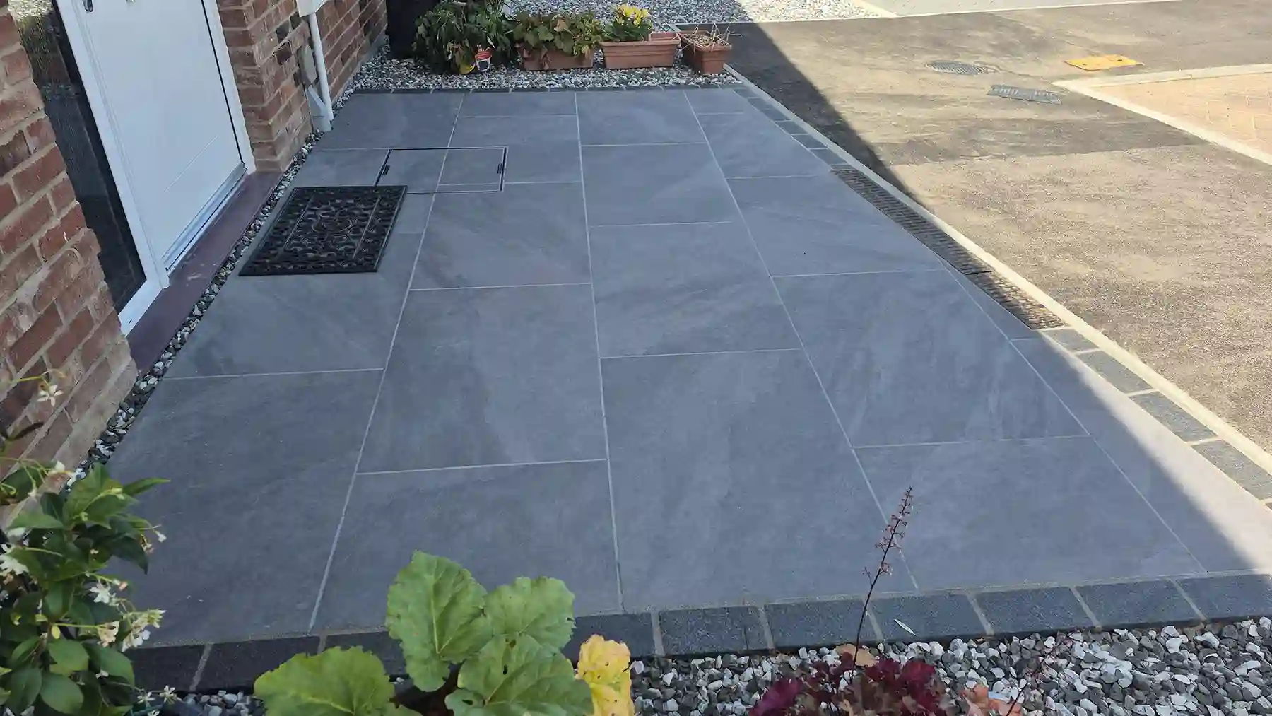 Angle view of a newly paved patio path made of smooth grey porcelain slabs bordered with dark granite edging bricks near potted plants and a house entry.