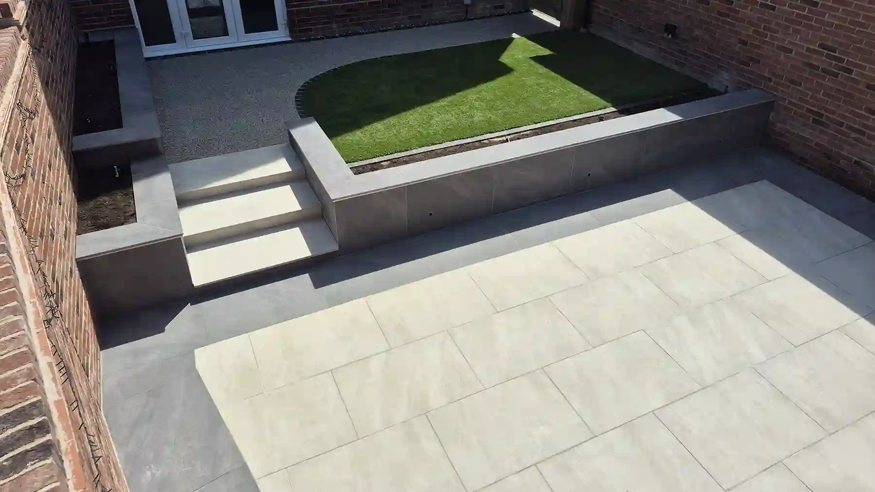 Sleek modern patio design combining resin surfacing, a lush artificial grass lawn, light beige paving slabs, and built-in dark grey planter walls.