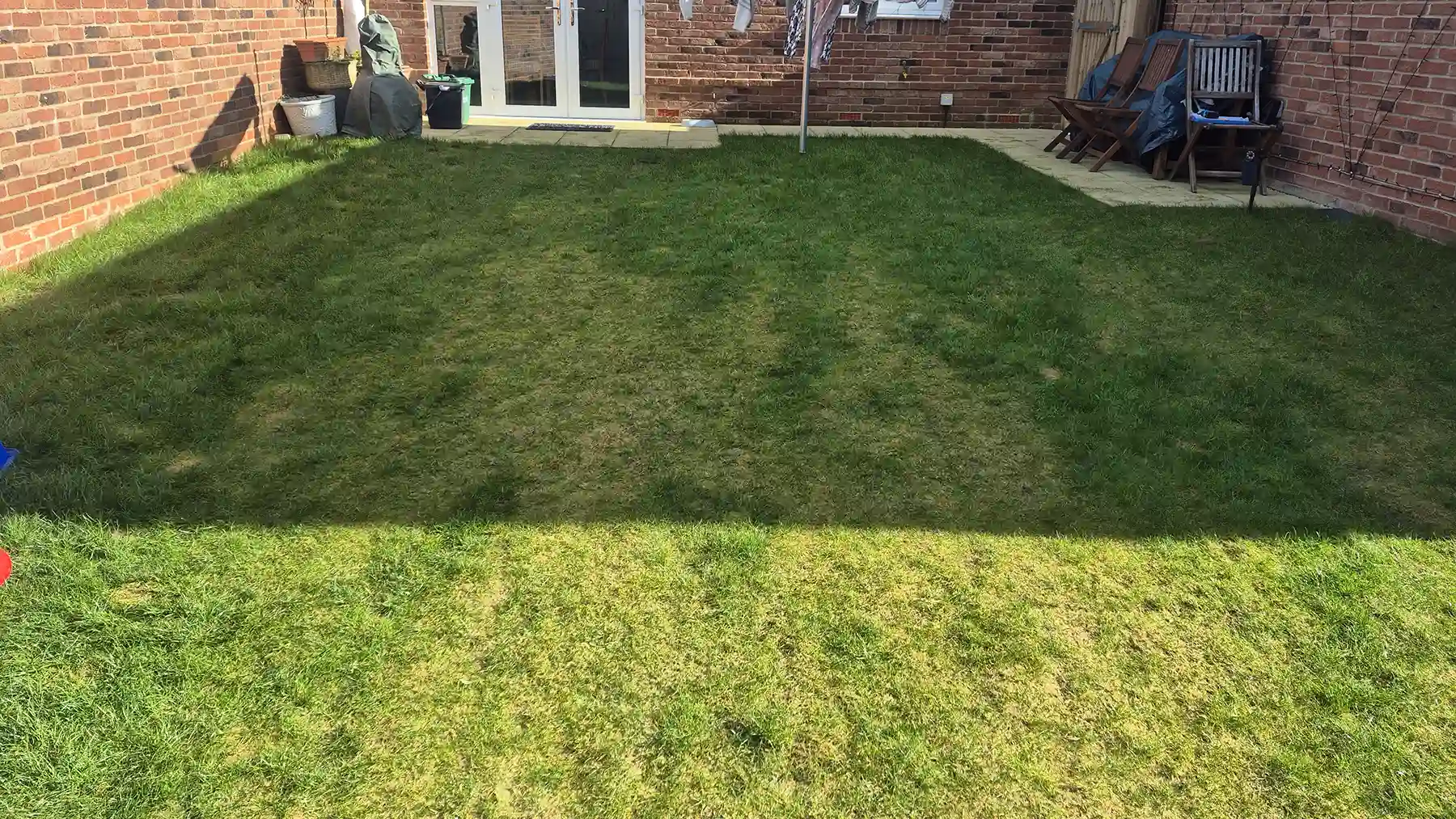 Overgrown green lawn in a new build garden before landscaping, enclosed by red brick walls, wooden fencing, and outdoor patio chairs.