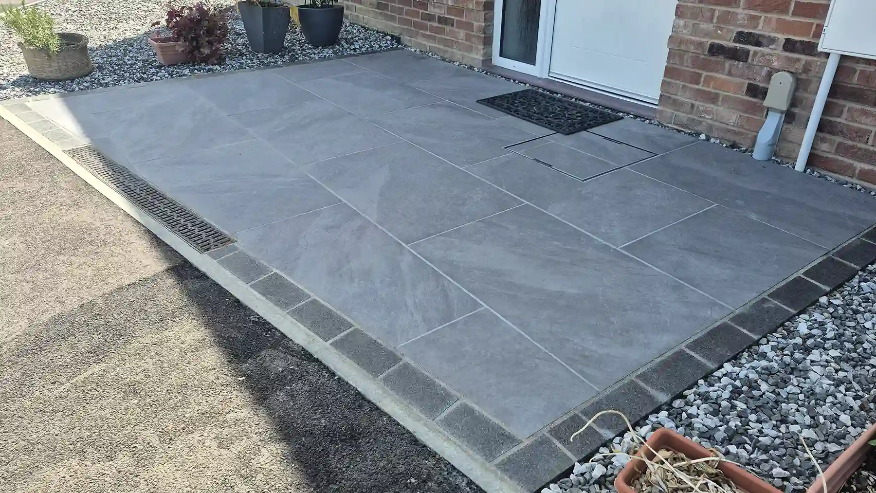 Ground-level view of smooth grey porcelain paving tiles framed by dark granite edging and gravel, incorporating a narrow drainage grate near the front doorstep.