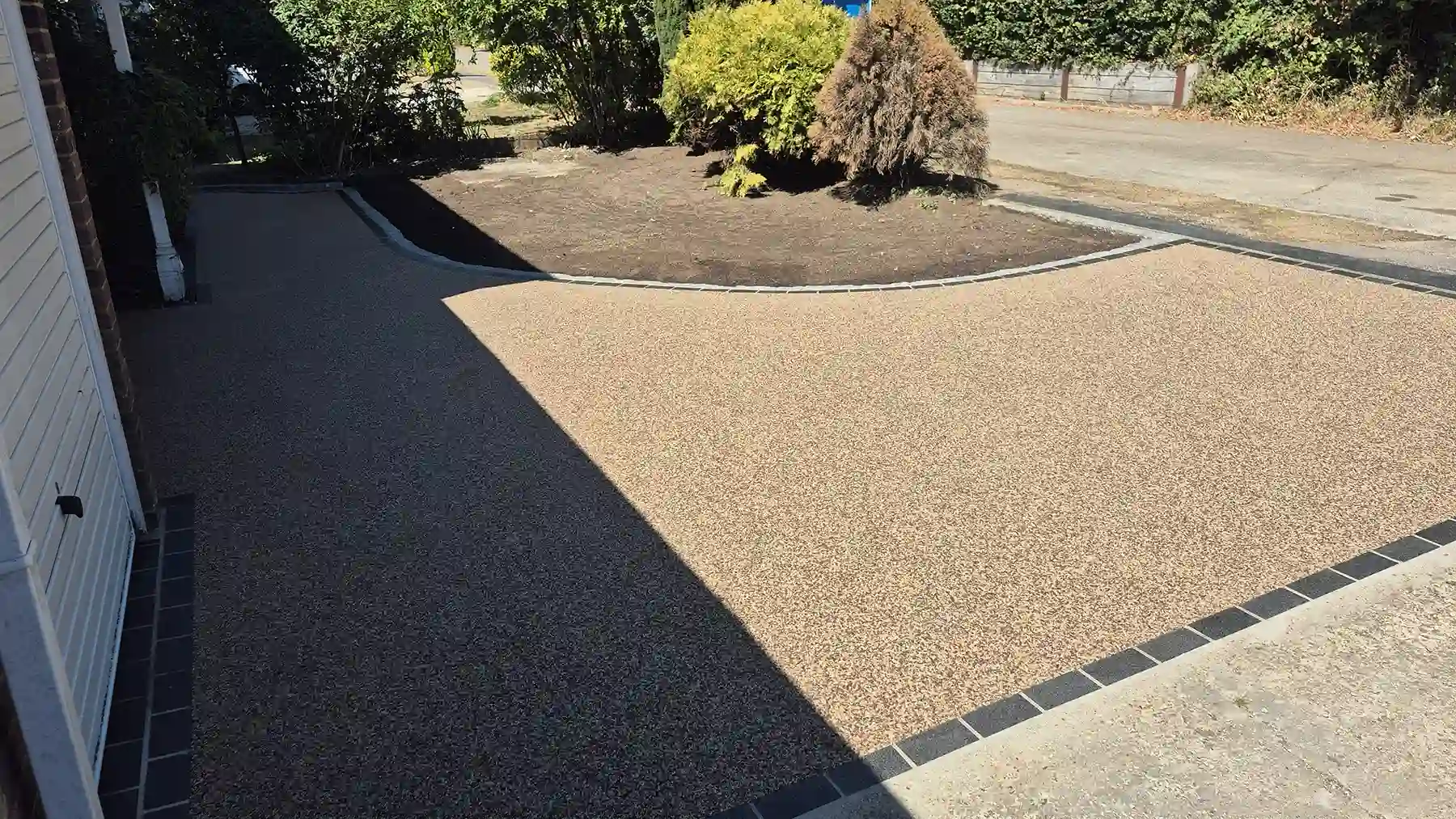 Golden resin bound driveway curving around a large landscaped flower bed, with established shrubs and contrasting dark granite edging.