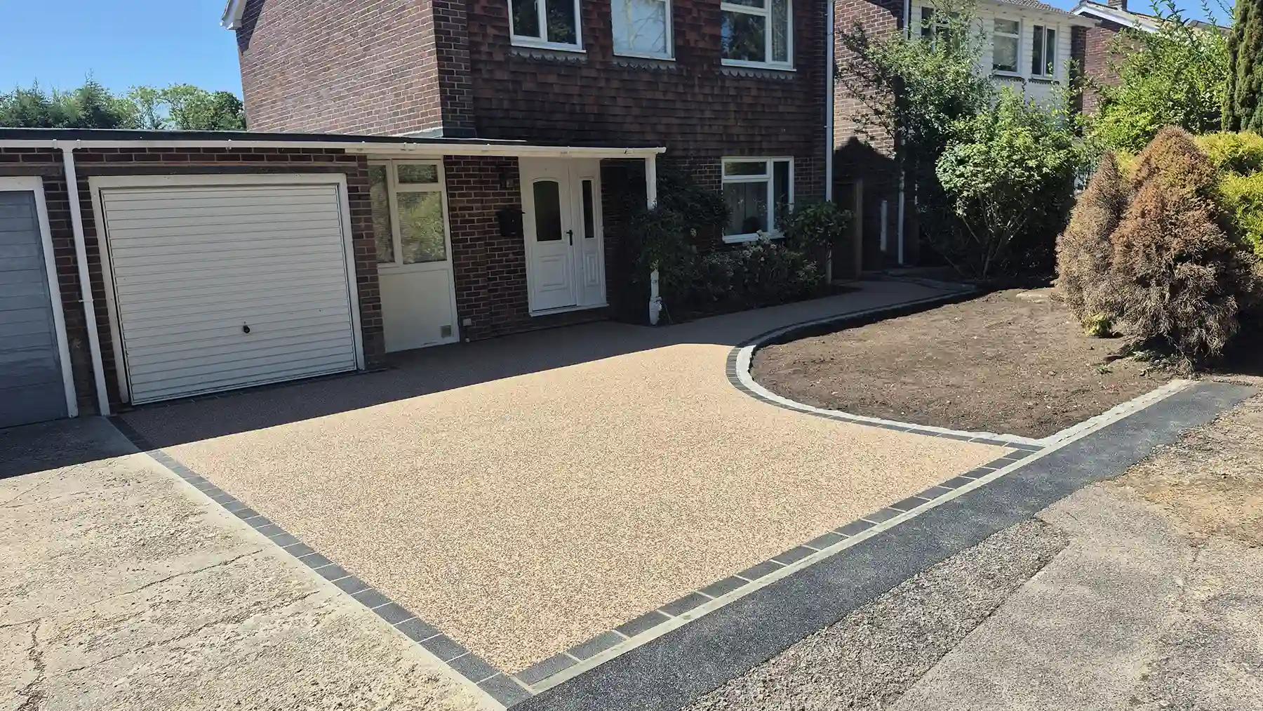Newly installed resin-bound driveway with a smooth golden-tan gravel surface, neat dark block borders, and space for garden planting next to a brick home and garage.
