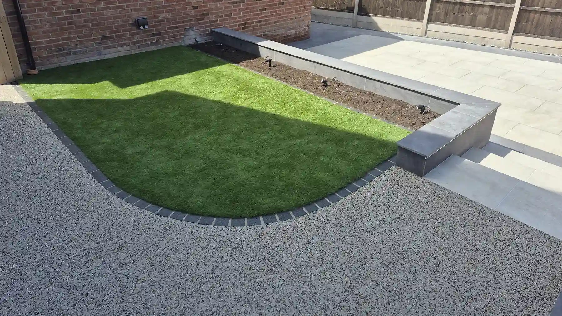 Curving resin bound path with artificial grass lawn with dark grey granite setts as the border, a raised planter box, and light porcelain patio steps in a landscaped garden.