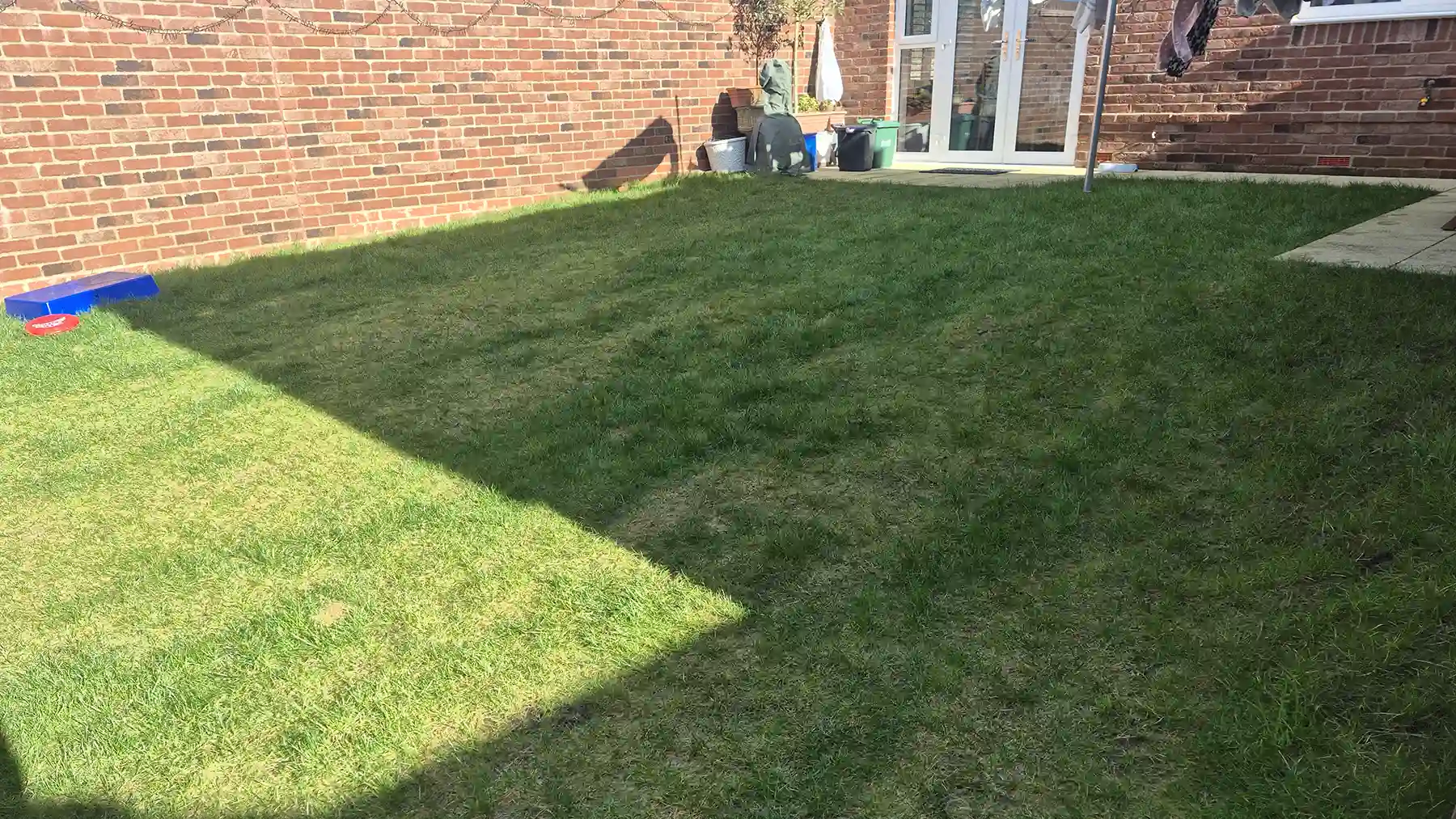 Broad view of an unlandscaped sloping back garden before landscaping, featuring a natural turf lawn behind a red brick home with white patio doors.